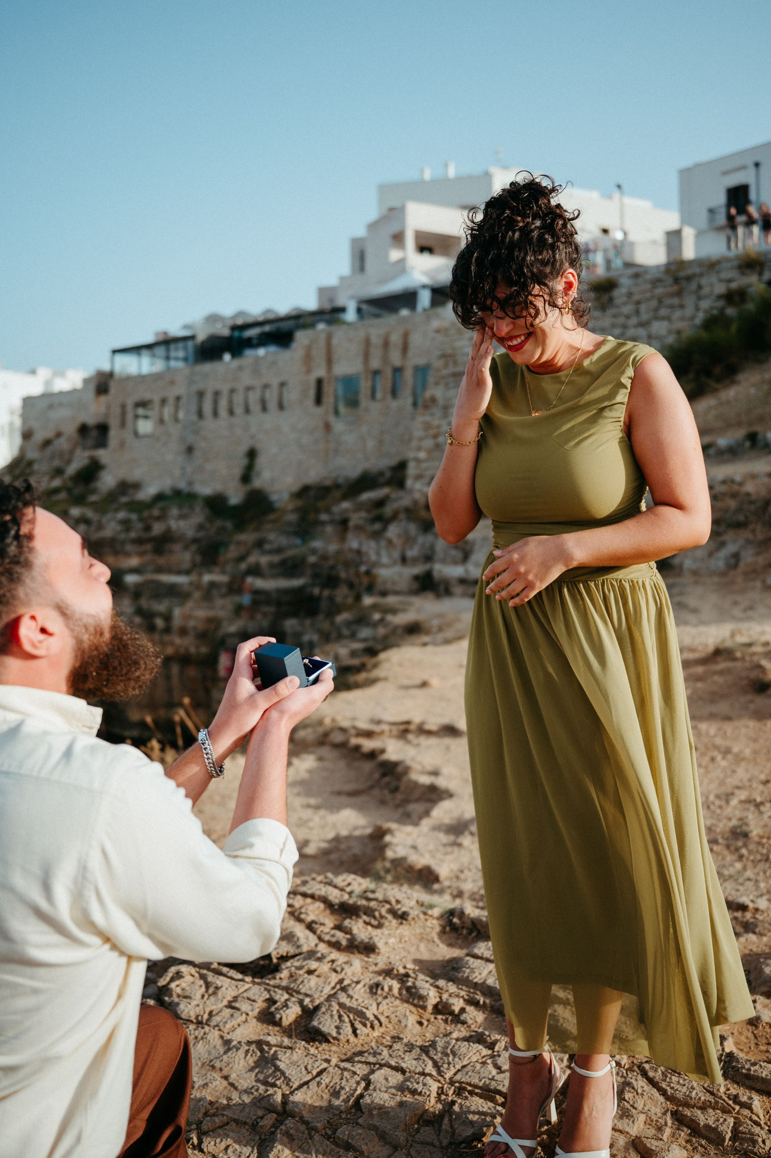 Man kneeling on rocky shore proposing to emotional woman in olive green dress with white buildings backdrop.