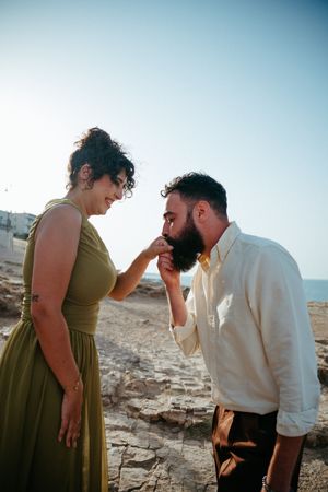 Bearded man kissing hand of smiling woman in olive dress on rocky Mediterranean coastline.