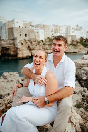 Laughing couple sitting on rocky cliffs with white Mediterranean buildings and turquoise sea in background.