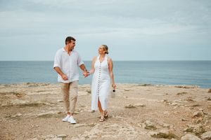 Couple in white outfits holding hands walking on rocky coastal cliff with ocean view.