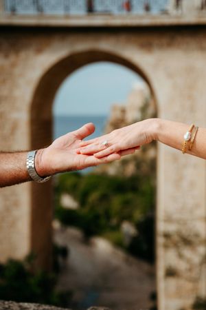 Close-up of couple holding hands with engagement ring, stone arch and sea in background.