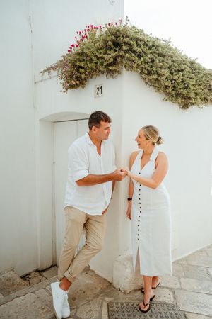 Couple holding hands and smiling outside white Mediterranean building with flowering vines above doorway.