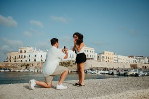 Man proposing to woman on one knee by a scenic coastal town, romantic marriage proposal outdoors.