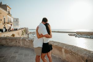 Couple embracing and kissing on a scenic Mediterranean coastal terrace with ocean views on a sunny day.
