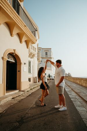 Couple dancing together on a scenic Mediterranean street with historic white buildings and ocean views at sunset.