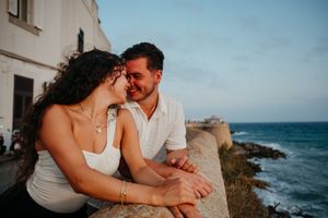 Romantic couple smiling at each other leaning on seaside wall at golden hour.