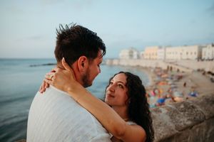 Romantic couple embracing by the seaside at dusk with a Mediterranean beach town in the background.