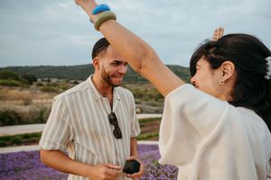 A smiling couple dances outdoors near a lavender field, with the woman raising her arm gracefully under a cloudy sky.