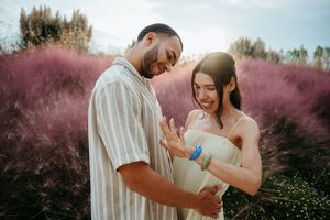 Couple standing in pink muhly grass field, man embracing pregnant woman admiring her baby bump outdoors.