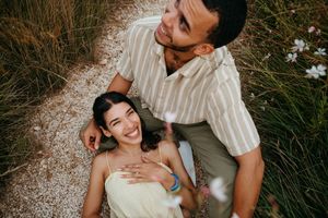 Couple sitting outdoors on gravel path surrounded by tall grass, woman smiling up at man who holds her affectionately.