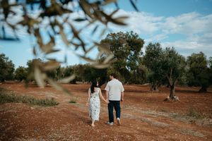 Romantic couple walking hand in hand through a scenic olive grove on red earth under a blue cloudy sky.
