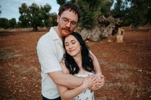 Couple embracing tenderly in a rustic olive grove with red soil, man hugging woman from behind as she closes her eyes.