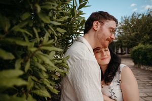 Loving couple sharing a tender moment beside lush green hedges, woman leaning back against mans chest outdoors.