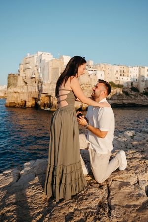 Man proposes to woman on rocky coastal cliffs with white buildings overlooking the sea.