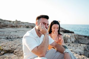 Emotional couple sitting on rocky coastal cliffs, both covering faces with hands, woman displaying engagement ring.