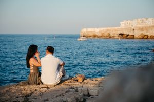 Couple sitting together on rocky cliffside overlooking blue Mediterranean sea with historic coastal town in background.