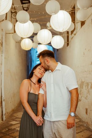 Couple holding hands and sharing a kiss under glowing white paper lanterns in a rustic alleyway at night.