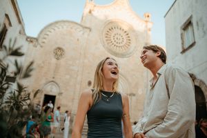 A laughing couple walks hand in hand in front of a historic cathedral in a sunny European town square.