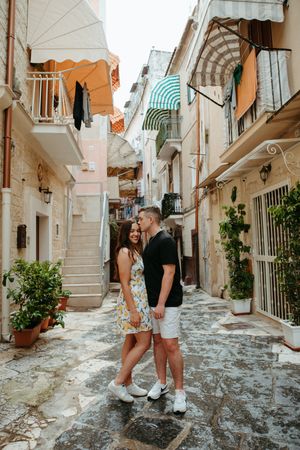 A romantic couple walks hand-in-hand through a charming narrow cobblestone alley lined with colorful Mediterranean buildings.