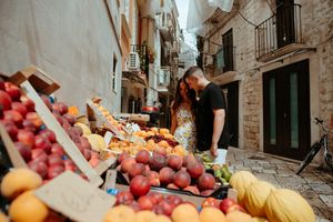 Couple browsing fresh fruit at an outdoor market in a charming European stone alleyway.
