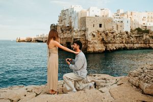 Man proposing to woman on rocky cliffside with stunning coastal Italian town backdrop.