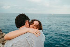 Couple sharing a tender embrace overlooking a stunning blue ocean on a cloudy day.