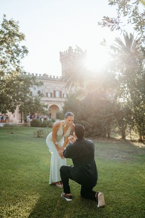 Man proposing on one knee to woman in white dress in sunlit castle garden.