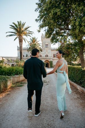 Couple holding hands walking toward a castle surrounded by palm trees and lush gardens.