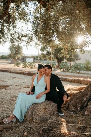 Couple sitting on hay bales under an olive tree at golden hour in a rustic countryside setting.