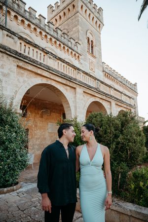 Elegant couple smiling at each other in front of a historic castle at dusk.