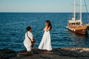 Man proposing to woman in white dress on rocky shore with sailboat in background on a sunny day.