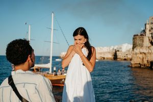 Man proposing to surprised woman in white dress by the sea with a sailboat and coastal cliffs in background.