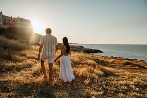 Couple holding hands watching golden sunset over Mediterranean sea on rocky coastal cliff.