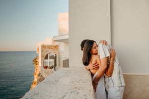 Couple embracing on a seaside terrace at sunset with white Mediterranean architecture and blue ocean views.