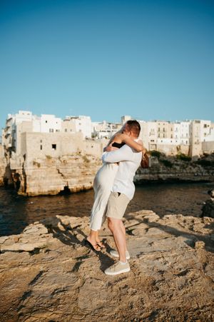Couple embracing on rocky cliffs with white Mediterranean buildings and clear blue sky in Polignano a Mare, Italy.