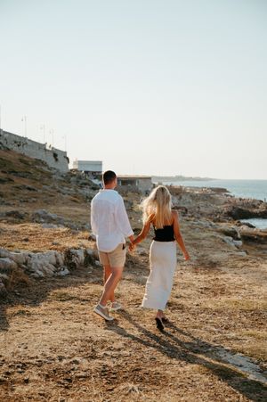 Couple holding hands walking along rocky coastal path at sunset, man in white shirt and woman in white skirt.