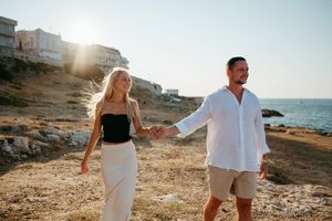 Couple holding hands walking along a sunny rocky coastline with the sea glowing behind them.