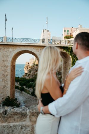Couple embracing while admiring a stone arch bridge overlooking the sea in a Mediterranean town.