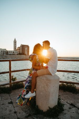 Romantic couple kissing on a stone pedestal at golden sunset with a historic cathedral and sea backdrop.