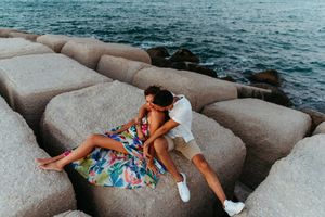 Romantic couple sitting together on large concrete boulders by the ocean, woman in colorful floral dress.