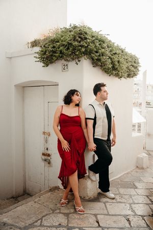 Stylish couple holding hands by white Mediterranean building, woman in red dress.