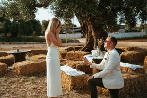 Man proposing on one knee to woman in white dress among hay bales under olive trees at sunset.