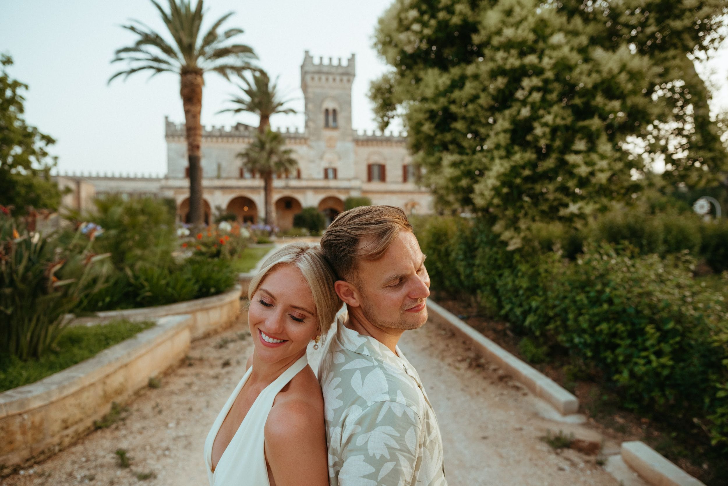 Happy couple back-to-back smiling before grand castle surrounded by palm trees.