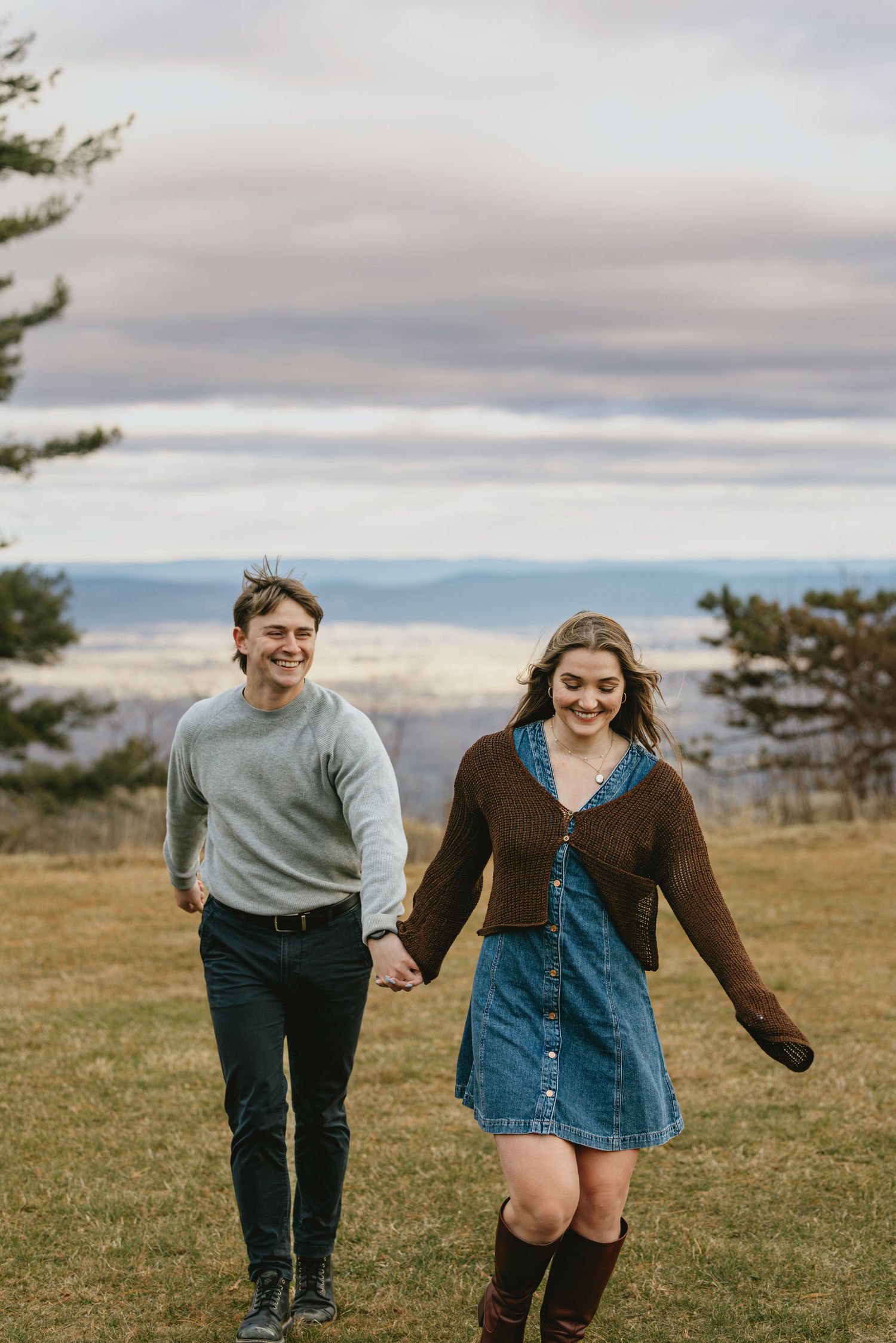 A happy young couple holds hands while walking through a grassy hilltop with scenic mountain views under a cloudy sky.