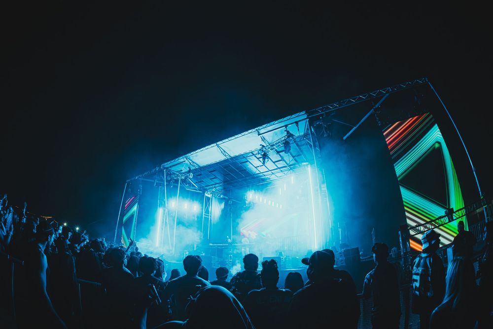 Crowd at an outdoor music festival with a glowing neon-lit stage and blue light effects at night.