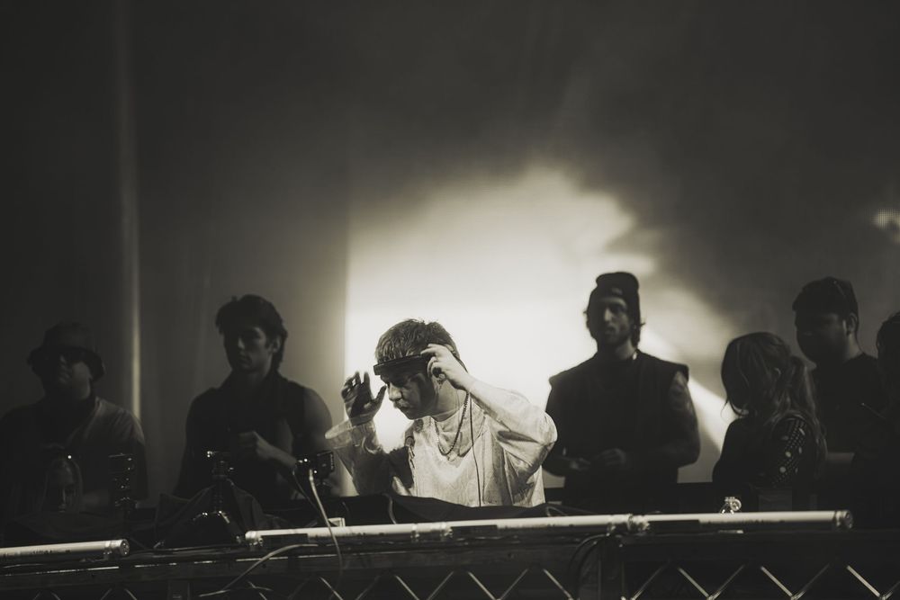 Black and white photo of a DJ performing on stage surrounded by people in a smoky, atmospheric concert venue.