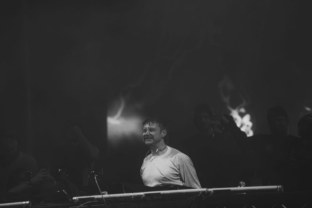 Black and white photo of a man smiling behind a DJ booth in a dark, atmospheric venue with dramatic lighting.