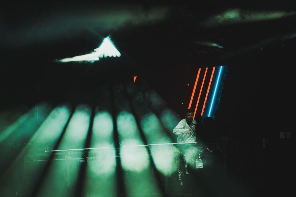 DJ performing on stage with vibrant neon light beams in green, blue, and orange illuminating a dark venue.