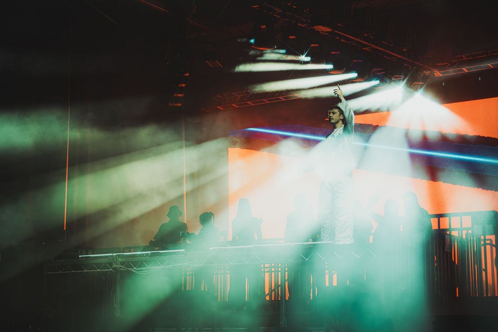 Concert stage with dramatic green and orange light beams cutting through fog in a dark venue.
