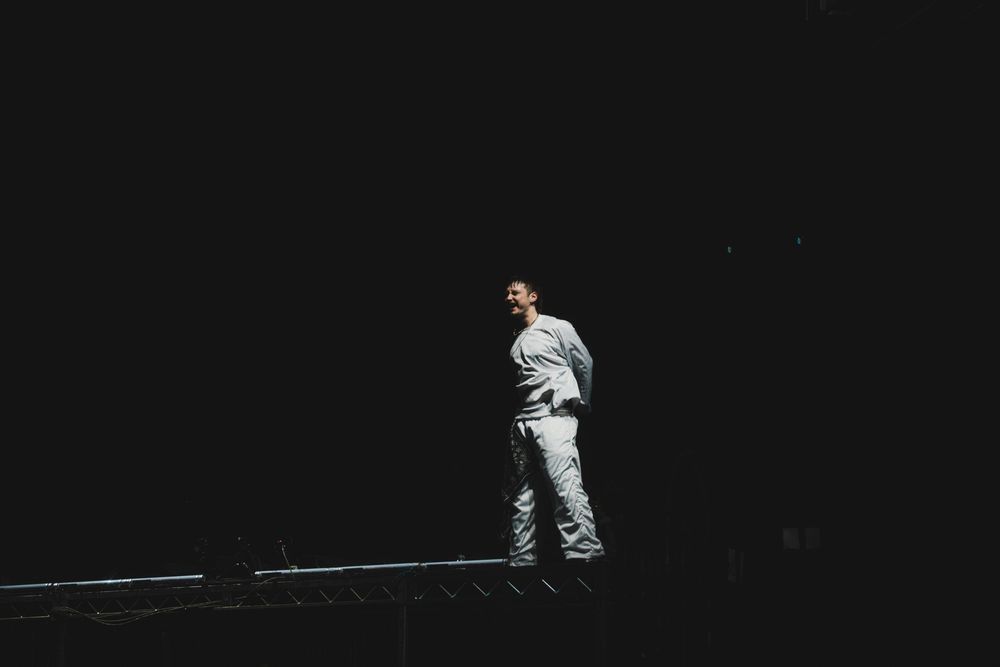 A lone man stands illuminated against a dark background, wearing light gray clothing in a dramatic, moody night scene.